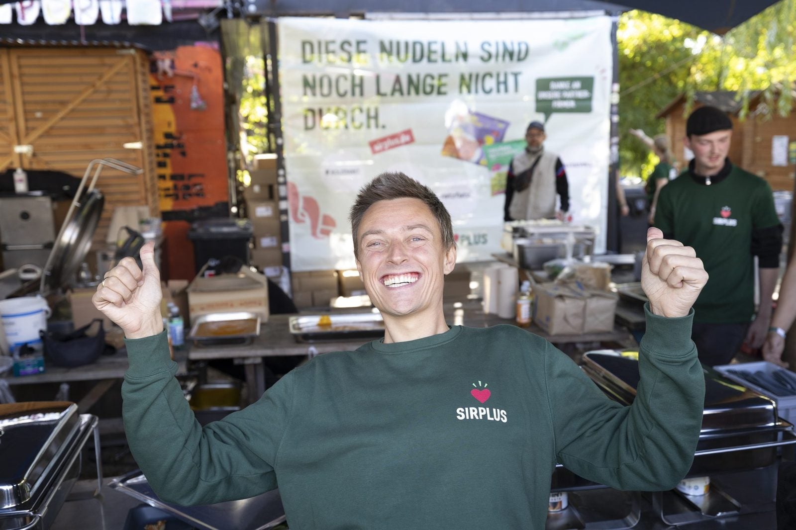 Mann im grünen SIRPLUS-Pullover jubelt mit erhobenen Armen vor vielen Besucher:innen am Holzmarkt