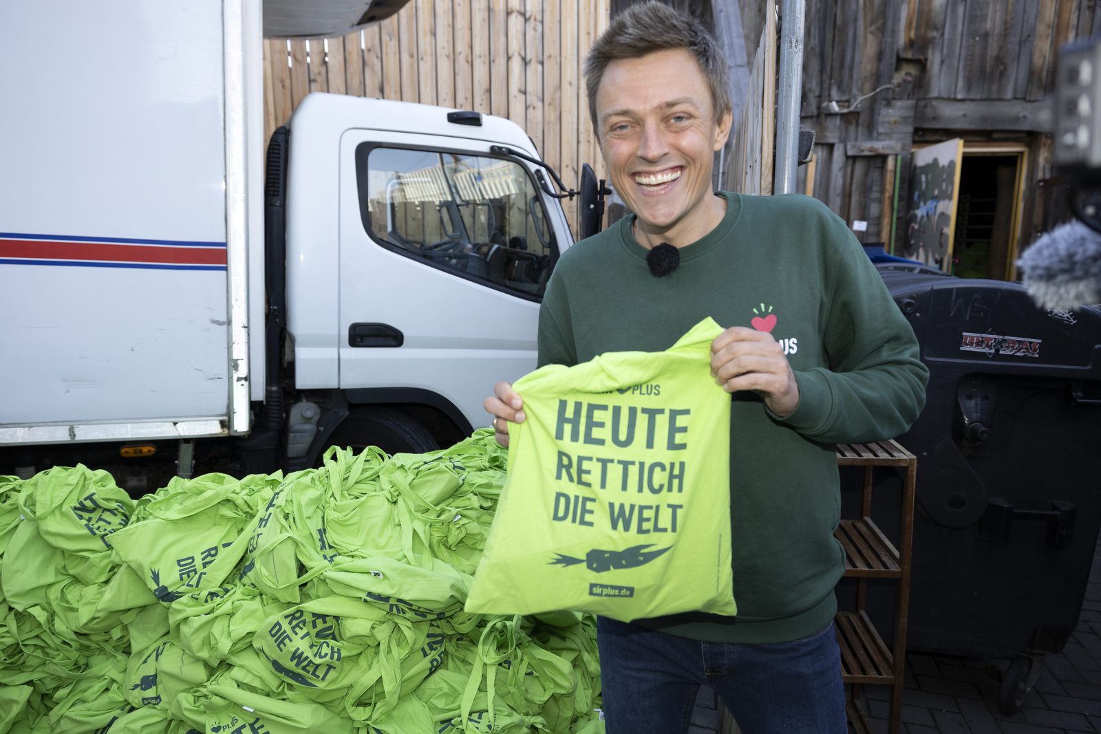 Mann im grünen SIRPLUS-Pullover mit gelben Stoffbeuteln vor einem Transporter