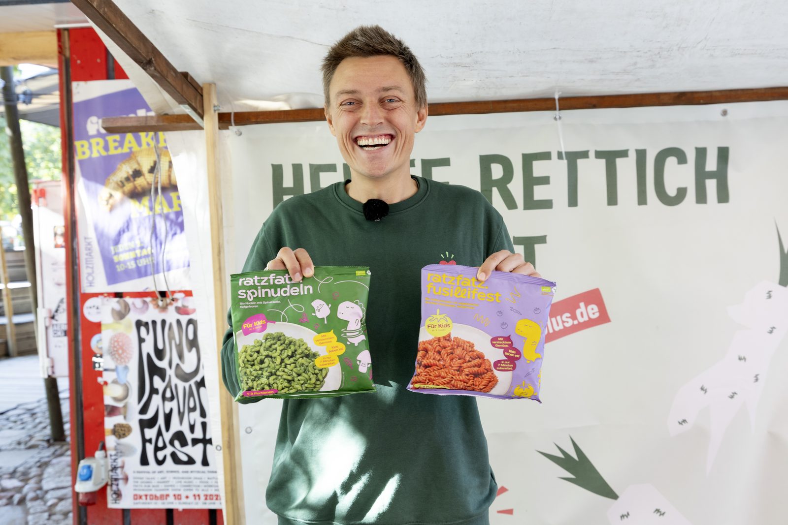Mann im SIRPLUS-Pullover hält Nudelpackungen vor einem Banner mit der Aufschrift Heute Rettich