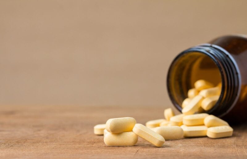 Gelbe Tabletten fallen aus einer umgekippten braunen Glasflasche auf einen Holztisch