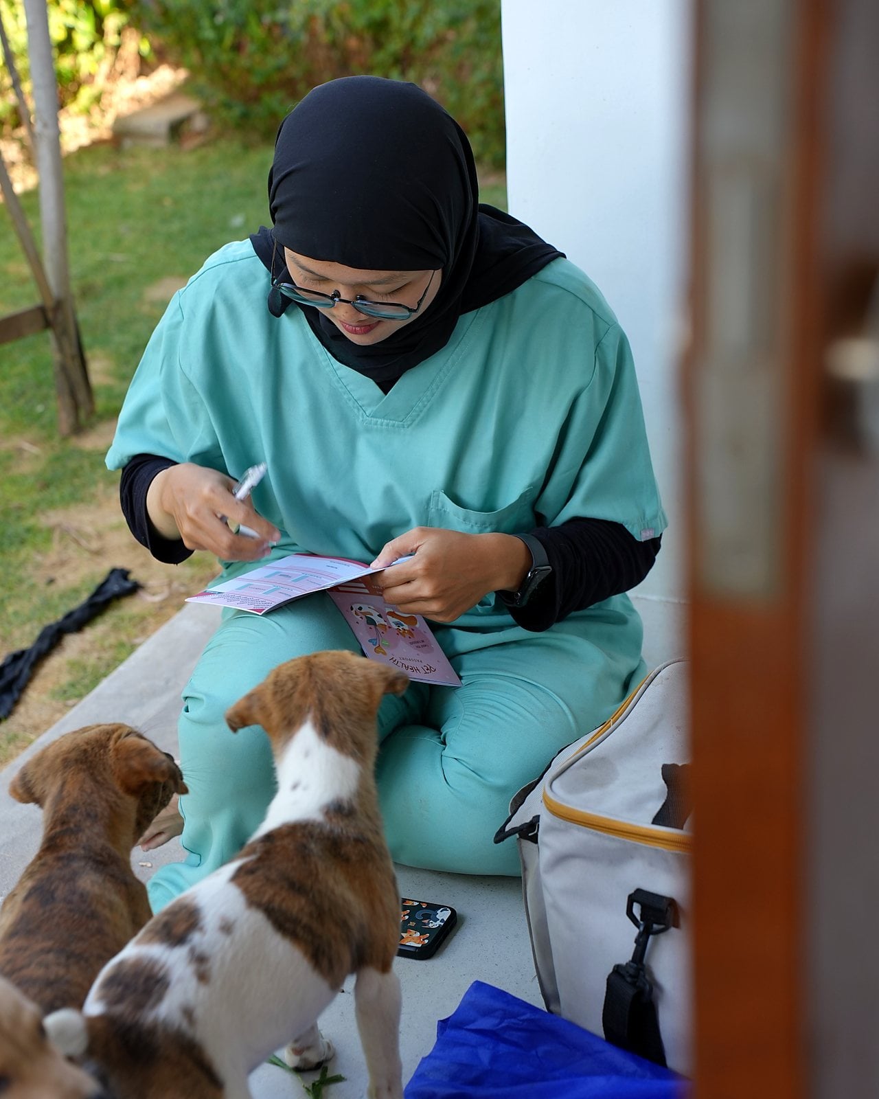 Tierärztin sitzt im Garten und füllt einen Impfpass aus, zwei Welpen stehen neben ihr