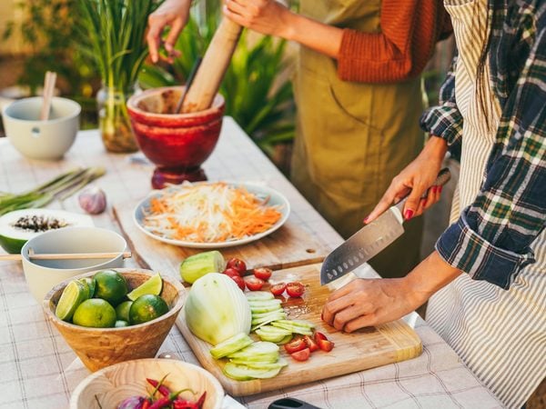 Hände schneiden Gemüse für einen Salat, daneben Mörser, Limetten und Kräuter