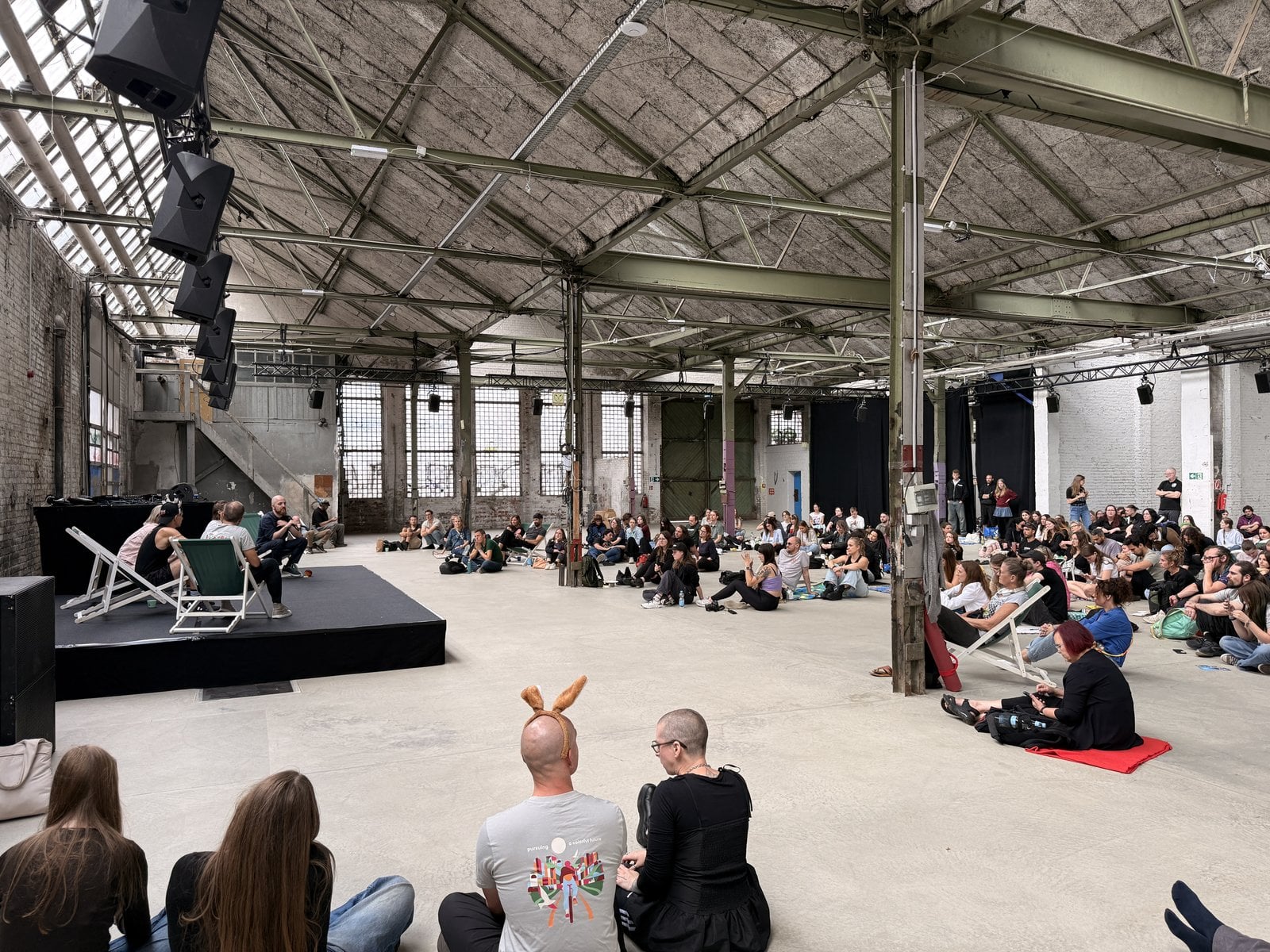 Publikum sitzt auf dem Hallenboden vor der Bühne und verfolgt die Panel-Diskussion