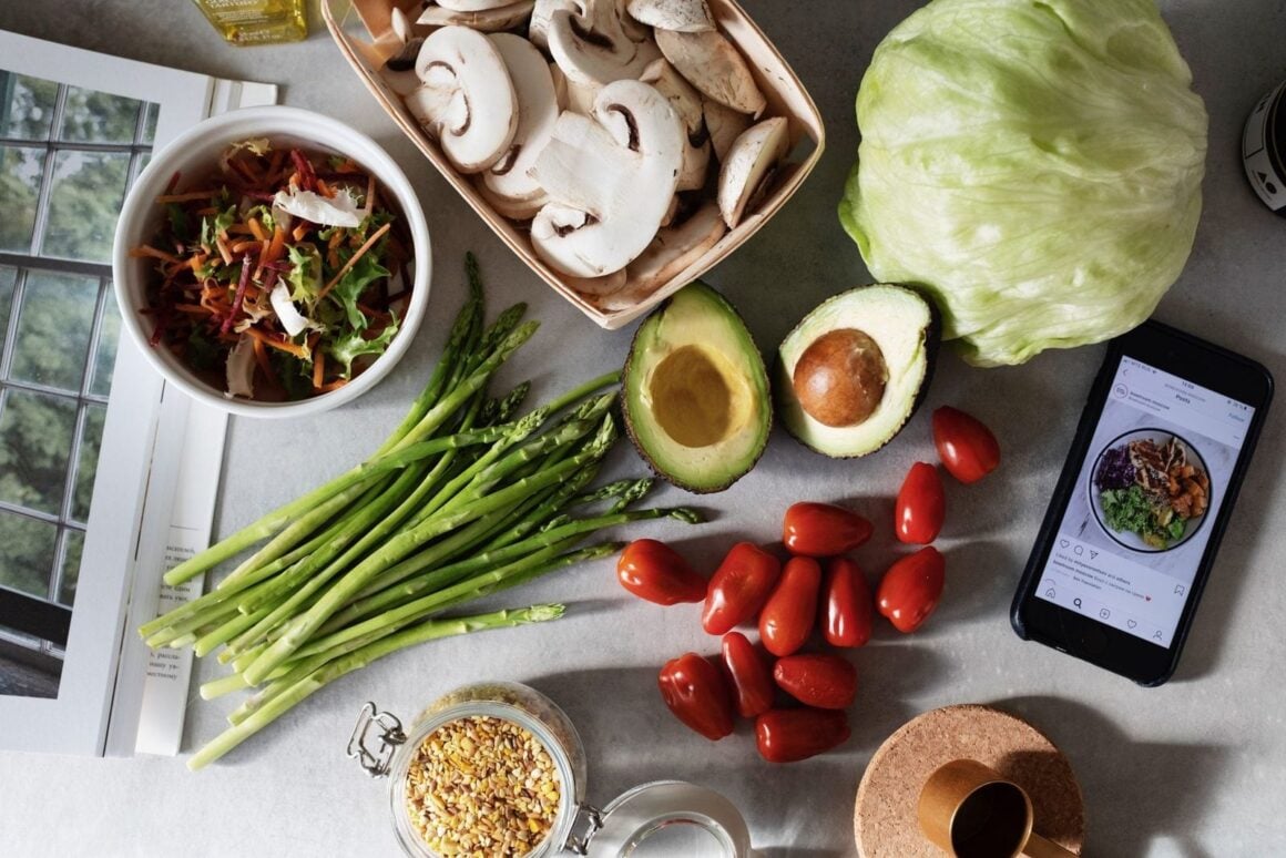 Frisches Gemüse von oben auf der Küchenarbeitsplatte: Spargel, Avocado, Kirschtomaten, Pilze, Salat und eine Salatschüssel neben einem Handy mit Rezept