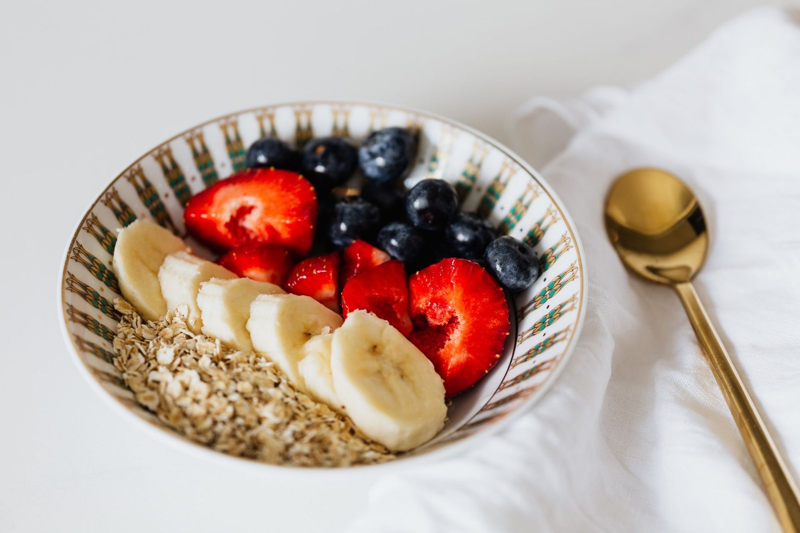 Bowl of oats topped with sliced banana, strawberries and blueberries, with a gold spoon on a white cloth