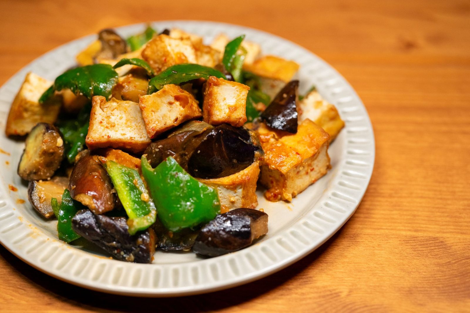 Plate of stir-fried tofu cubes with eggplant and green bell peppers on a wooden table