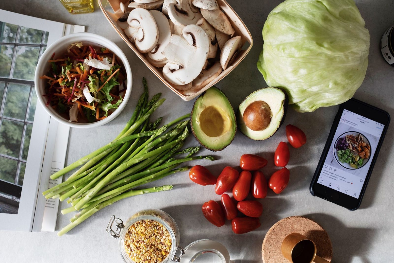 Top view of fresh vegetables on a kitchen counter: asparagus, avocado, cherry tomatoes, mushrooms, lettuce and a salad bowl next to a phone showing a recipe