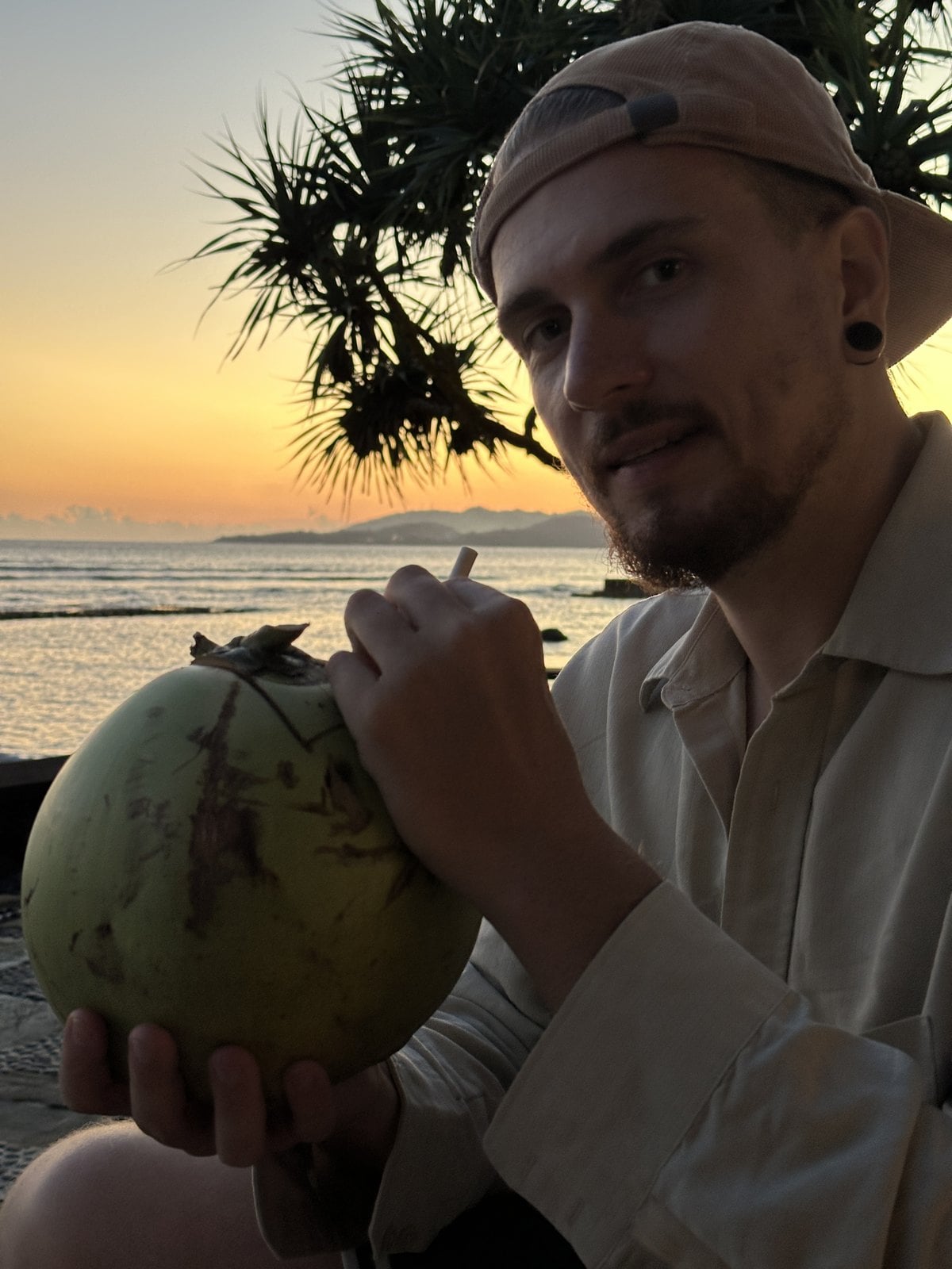 Yannick hält bei Sonnenuntergang am Strand eine frisch geöffnete grüne Kokosnuss mit Strohhalm