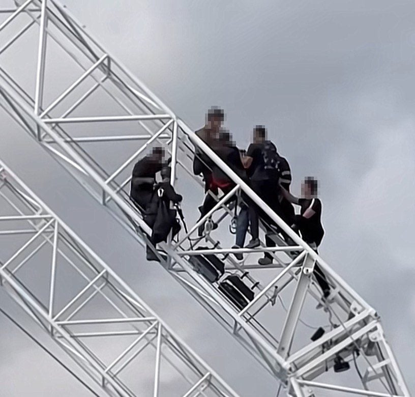 Mehrere Personen stehen dicht beieinander auf einer weißen Stahl-Gitterkonstruktion vor bewölktem Himmel