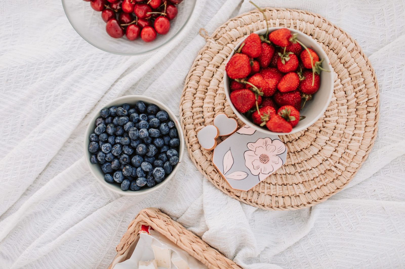 Draufsicht auf einen hellen Tisch mit weißem Leinentuch: eine Schale Blaubeeren, eine Schale Erdbeeren auf einem geflochtenen Untersetzer und am oberen Rand eine Schale Kirschen.