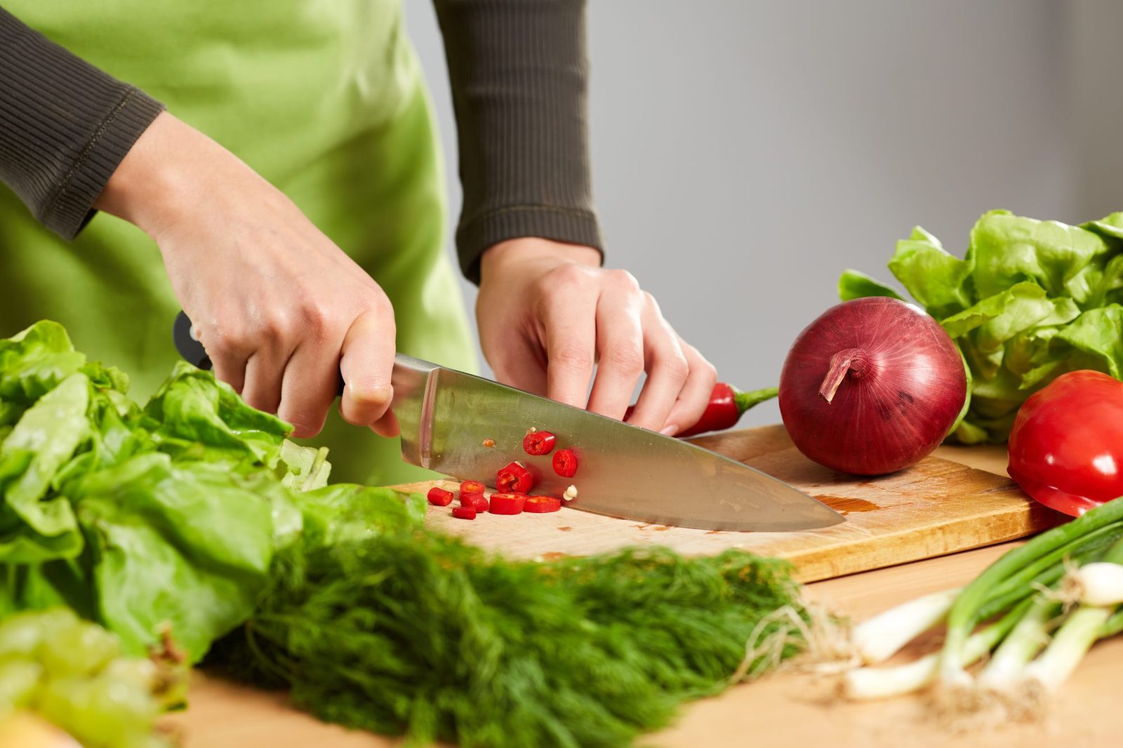 Zwei Hände schneiden eine rote Chili auf einem hellen Holzbrett, umgeben von Kopfsalat, Dill, einer roten Zwiebel, roter Paprika und Frühlingszwiebeln.