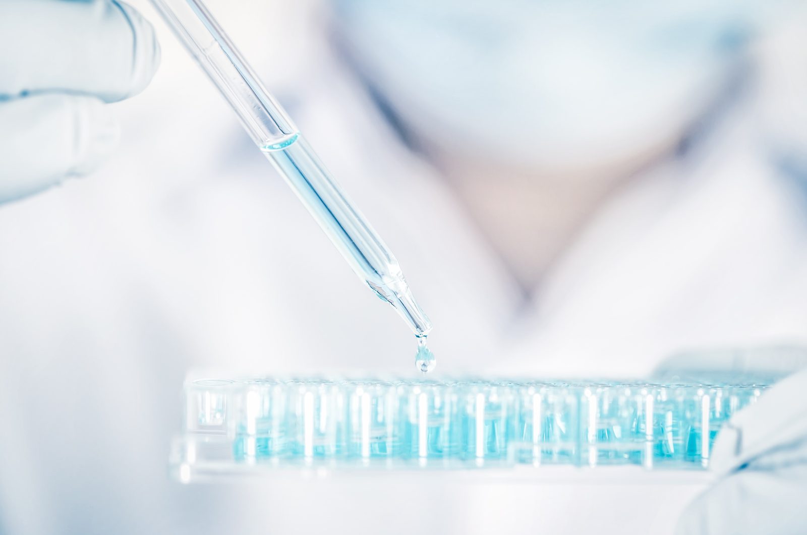 Eine Pipette gibt blaue Flüssigkeit in eine Mikrotiterplatte, dahinter unscharf eine Person im Laborkittel