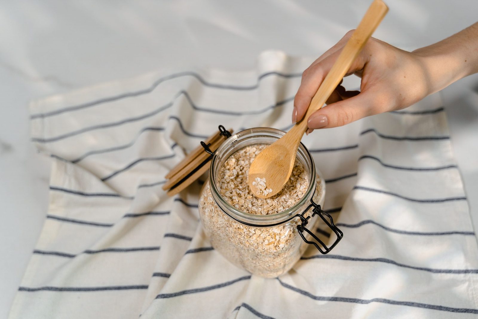 Eine Hand schöpft mit einem Holzlöffel Haferflocken aus einem Bügelglas, das auf einem weiß-grau gestreiften Leinentuch steht.