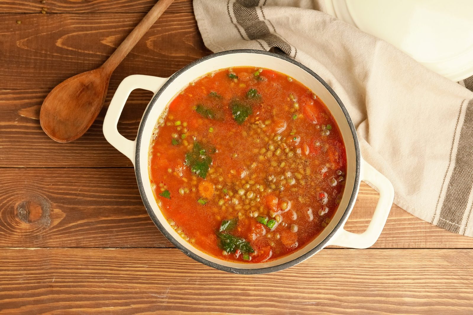 Draufsicht auf einen cremefarbenen Emailtopf mit Linsensuppe in Tomatenbrühe mit Karotten und Kräutern auf einem Holztisch, daneben ein Holzlöffel und ein Leinentuch.