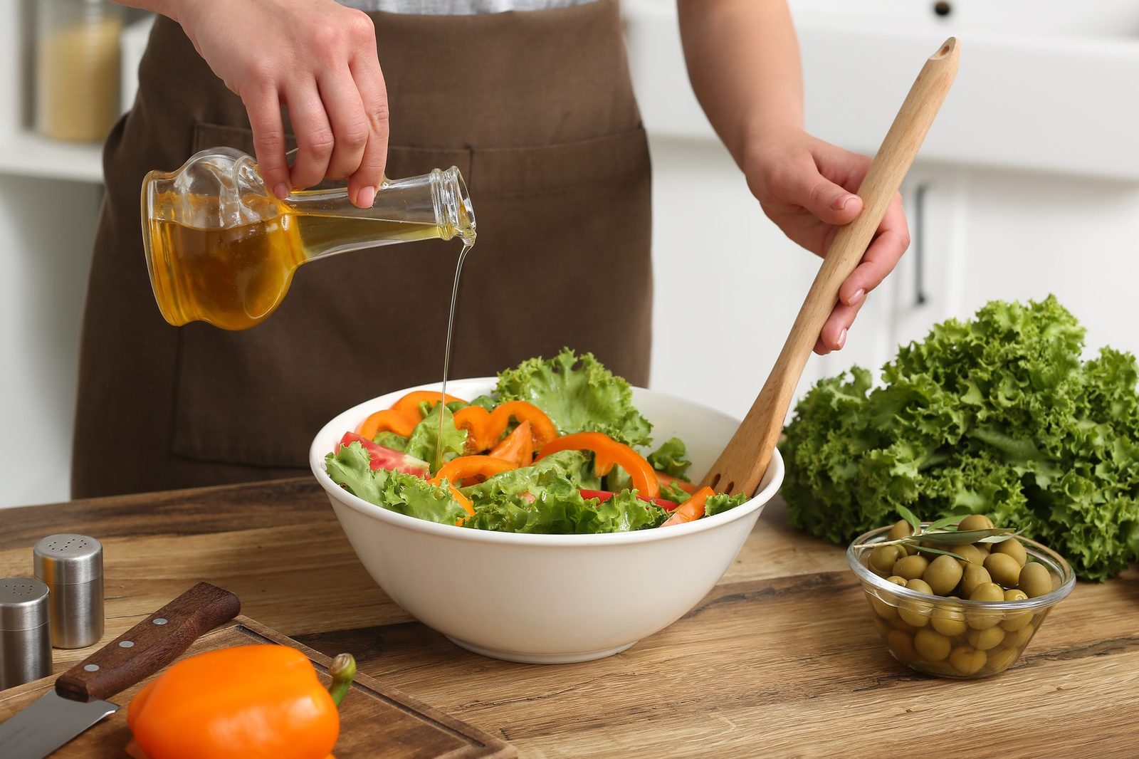 Eine Person mit brauner Schürze gießt Olivenöl aus einer Glasflasche über einen Salat aus Blattsalat und Paprikastreifen in einer weißen Schüssel und rührt mit einem Holzlöffel um; auf der Arbeitsfläche liegen Paprika, Oliven und Salatblätter.