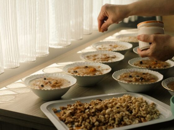 Zwei Hände streuen Toppings aus einem kleinen Glas über mehrere Schalen Porridge, die auf einem hellen Tisch vor einem lichtdurchfluteten Vorhang stehen; im Vordergrund ein Blech mit gehackten Walnüssen.