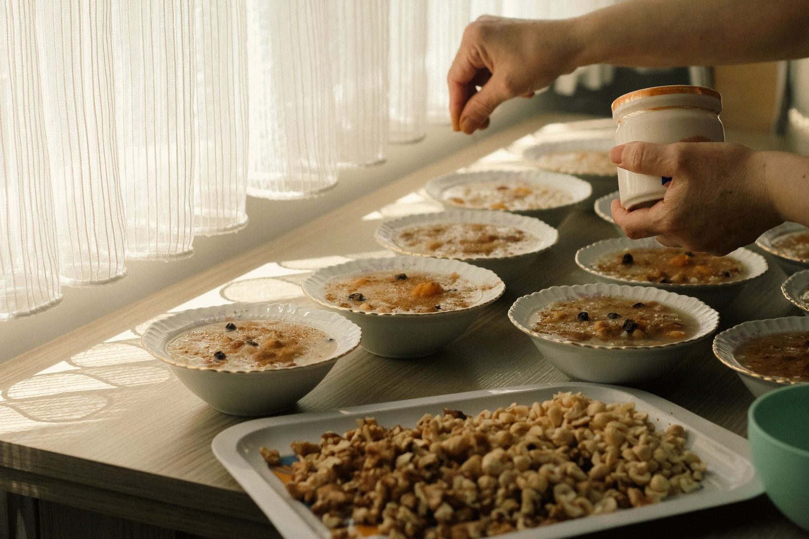 Zwei Hände streuen Toppings aus einem kleinen Glas über mehrere Schalen Porridge, die auf einem hellen Tisch vor einem lichtdurchfluteten Vorhang stehen; im Vordergrund ein Blech mit gehackten Walnüssen.