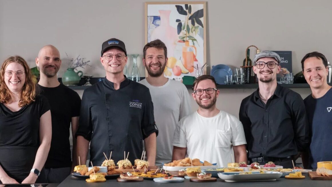 Sebastian Copien in schwarzer Kochjacke steht mit dem Team von 7hauben hinter einem Tisch mit Fingerfood