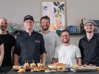 Sebastian Copien in schwarzer Kochjacke steht mit dem Team von 7hauben hinter einem Tisch mit Fingerfood