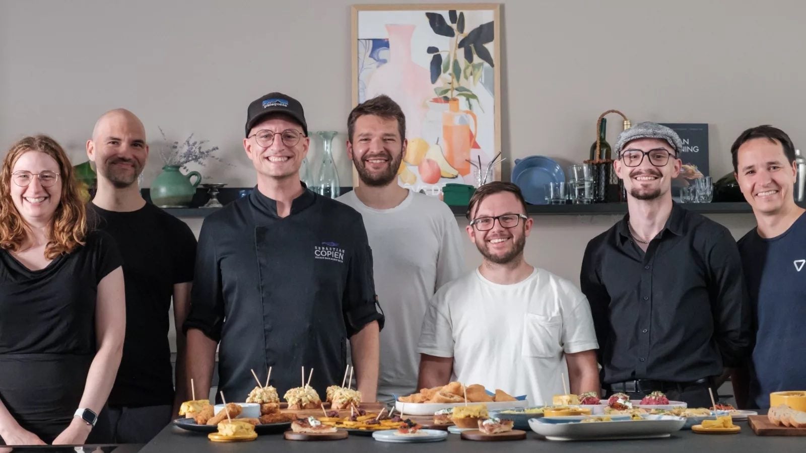 Sebastian Copien in schwarzer Kochjacke steht mit dem Team von 7hauben hinter einem Tisch mit Fingerfood