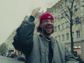 FAYAN mit roter Cap und Bomberjacke lacht mit erhobener Hand auf einer Berliner Straße