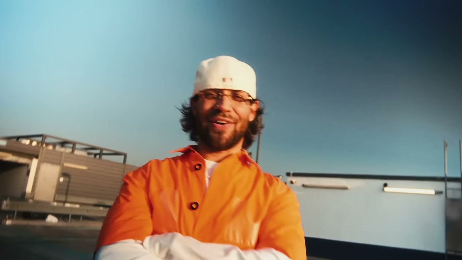 FAYAN in oranger Jacke und weißer Cap lacht auf einem Dach vor blauem Himmel
