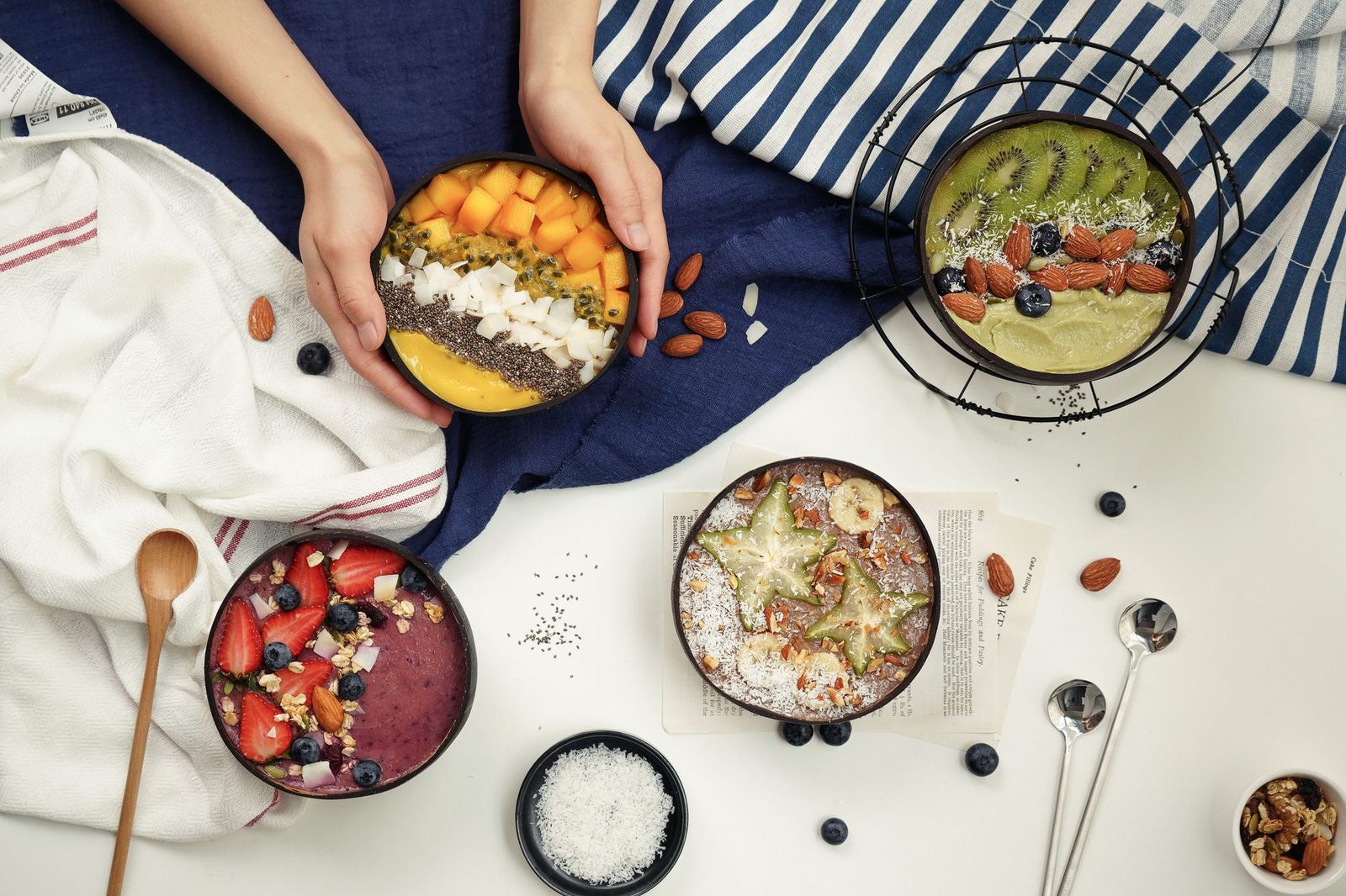Gedeckter Frühstückstisch von oben: Hände halten eine Smoothie-Bowl mit Mango, Kokos und Chiasamen, daneben Bowls mit Beeren, Kiwi und Sternfrucht, verstreute Blaubeeren, Mandeln und ein Schälchen Kokosraspel auf weißem Tisch mit blauen und gestreiften Tüchern.