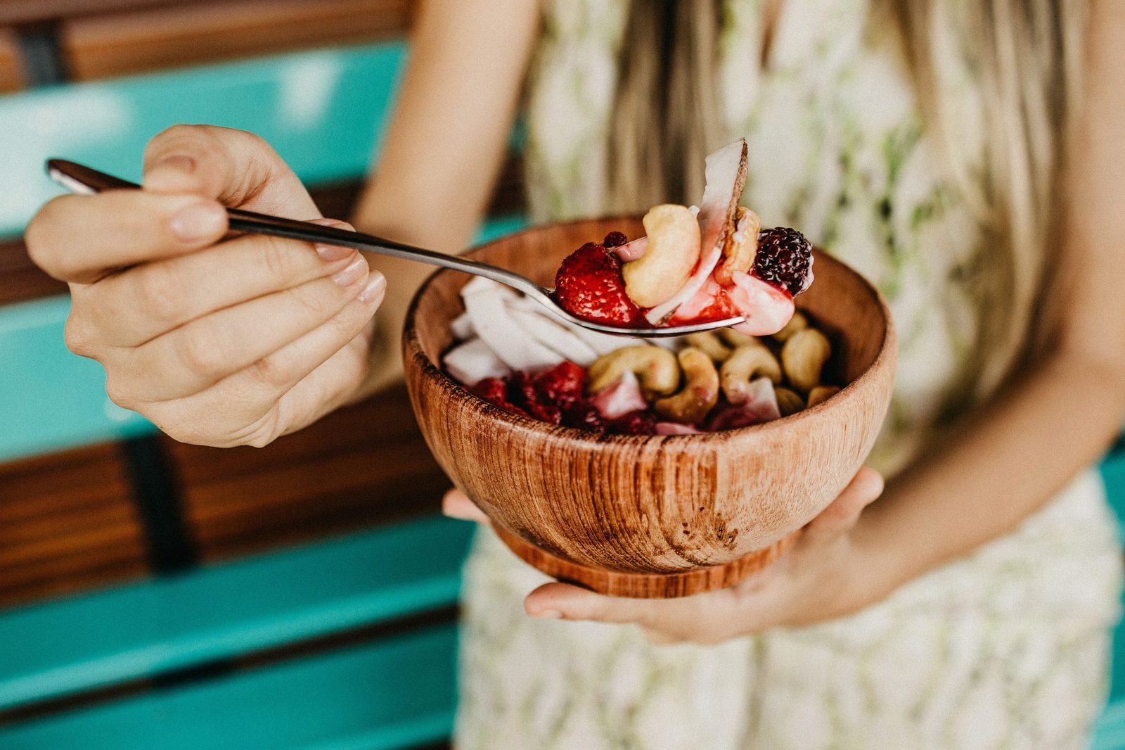 Frau im hellen Sommerkleid hält eine Holzschale mit Açaí-Bowl und nimmt mit dem Löffel Erdbeeren, Cashews und Kokosstücke, im Hintergrund eine türkisfarbene Holzbank.
