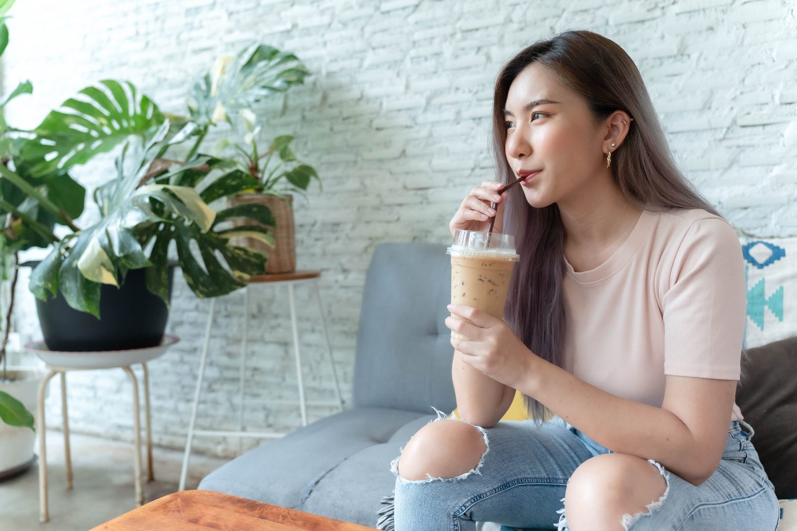 Junge Frau im rosa T-Shirt sitzt morgens auf dem Sofa vor einer weißen Backsteinwand mit Monstera und trinkt einen hellbraunen Kaffee-Shake mit Strohhalm aus einem Becher.