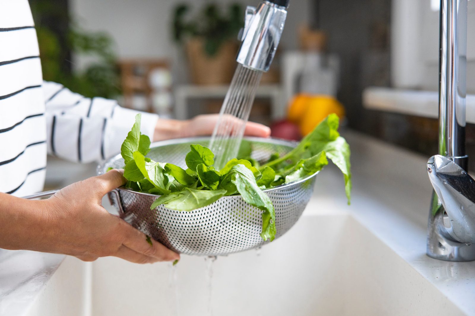 Zwei Hände halten ein Edelstahlsieb voller frischer grüner Blätter unter den Wasserhahn einer hellen Küchenspüle.