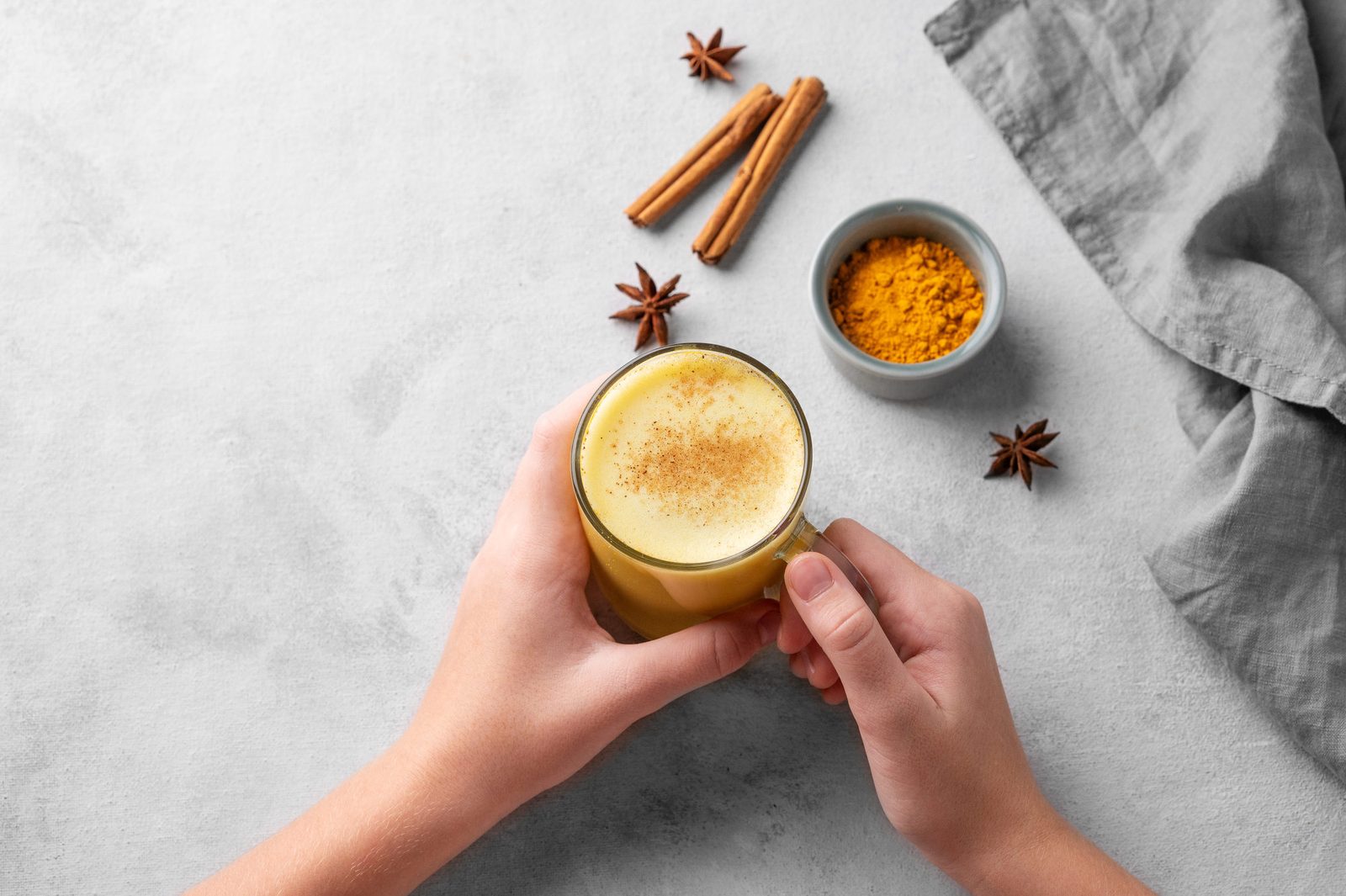 Zwei Hände halten von oben ein Glas mit goldgelber Kurkuma-Milch mit Zimt bestäubt, daneben Zimtstangen, Sternanis, ein Schälchen Kurkumapulver und ein graues Leinentuch auf heller Steinplatte.