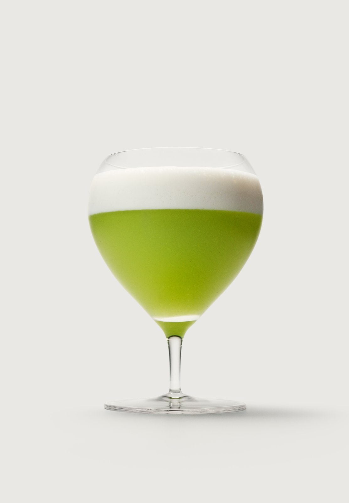 Ein grüner Drink mit weißem Schaum in einem Stielglas vor hellgrauem Hintergrund