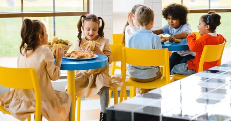 Kinder essen an bunten Tischen in einer Schulmensa zu Mittag