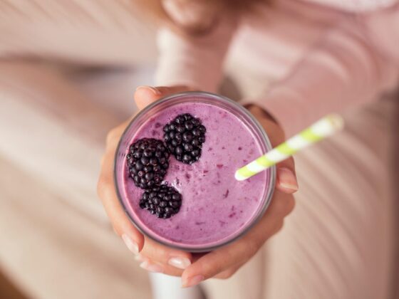 Zwei Hände halten von oben ein Glas mit violettem Beeren-Smoothie, obenauf drei Brombeeren und ein gelb-weiß gestreifter Strohhalm; im unscharfen Hintergrund ein heller Strickpullover.