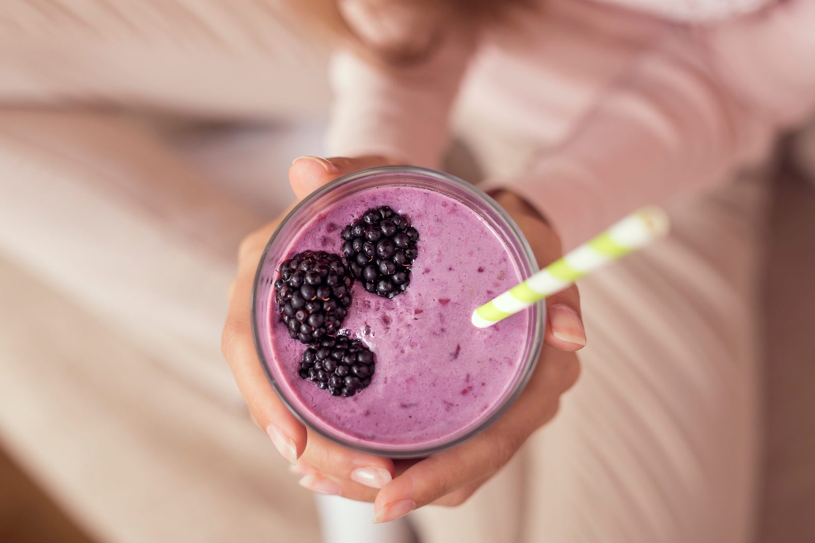 Zwei Hände halten von oben ein Glas mit violettem Beeren-Smoothie, obenauf drei Brombeeren und ein gelb-weiß gestreifter Strohhalm; im unscharfen Hintergrund ein heller Strickpullover.