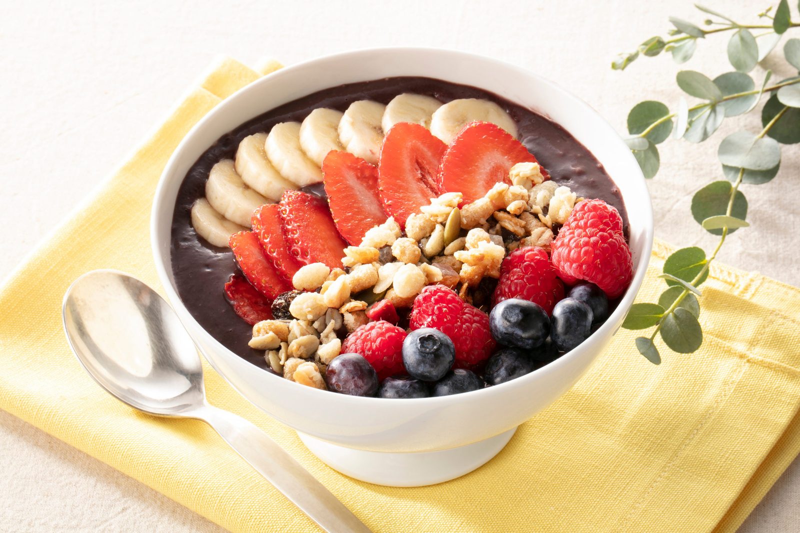 Weiße Schale mit dunkelvioletter Açaí-Smoothie-Bowl, belegt mit Bananenscheiben, Erdbeeren, Himbeeren, Blaubeeren und Nuss-Kerne-Mischung, auf einer gelben Serviette mit Löffel, daneben ein Eukalyptuszweig auf hellem Untergrund.