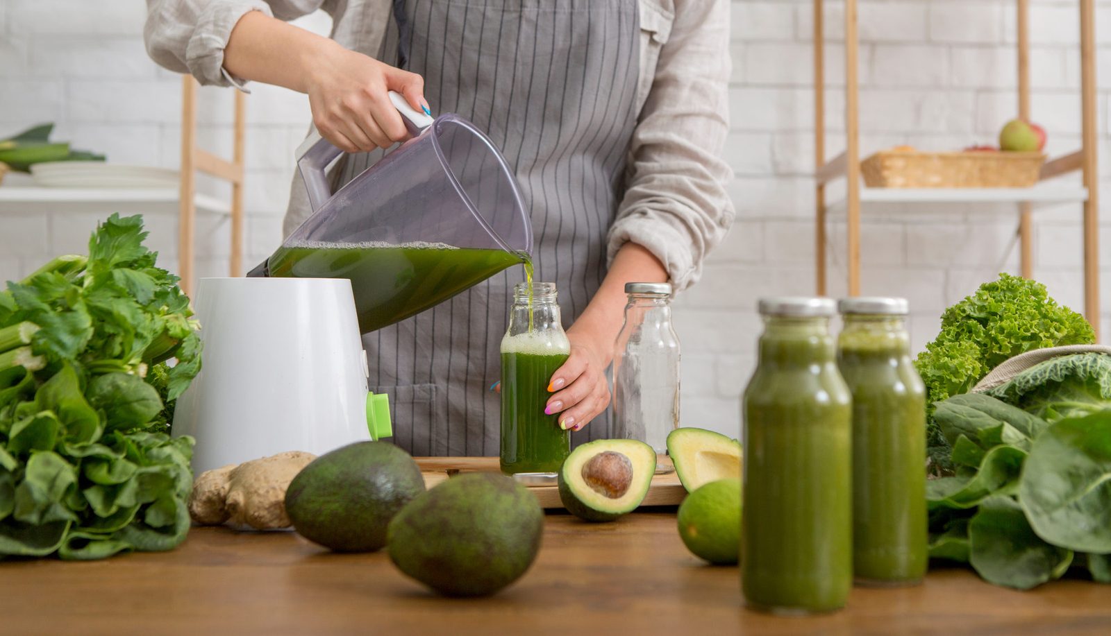 Person in gestreifter Leinenschürze gießt grünen Smoothie aus dem Mixbecher in eine Glasflasche; auf dem Holztisch stehen weitere gefüllte Flaschen, Avocados, Ingwer, Salat und Spinat, im Hintergrund ein helles Küchenregal.