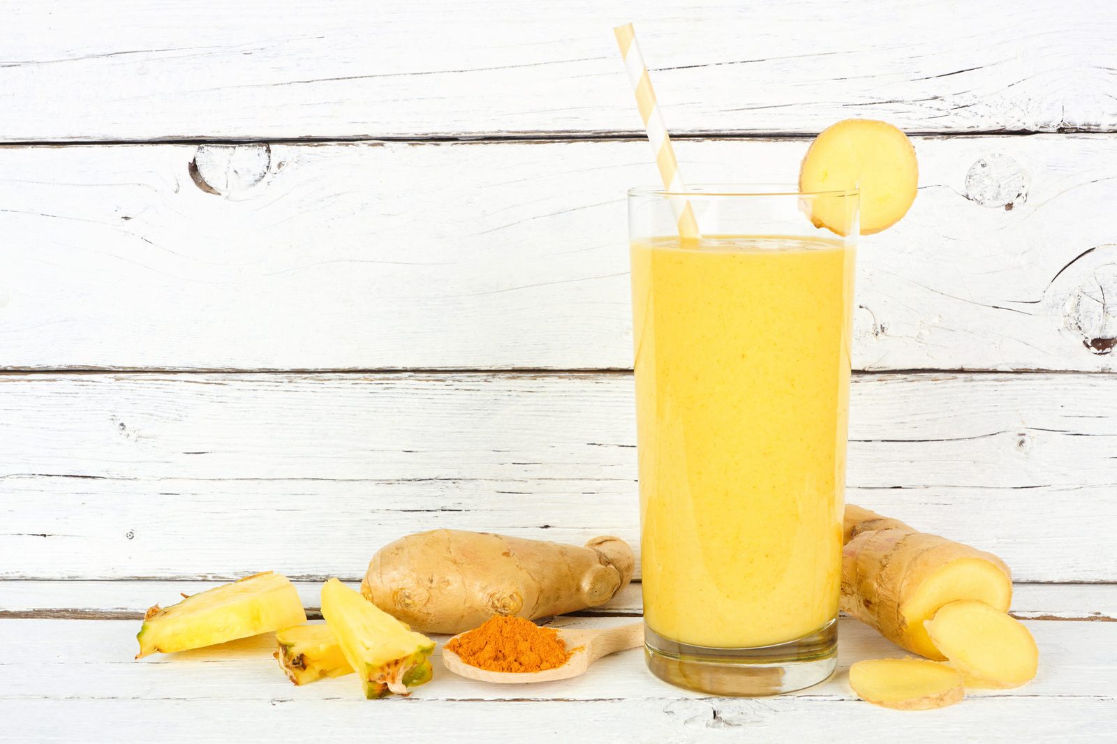 Hohes Glas mit gelbem Ananas-Kurkuma-Smoothie und gestreiftem Strohhalm, am Rand eine Ingwerscheibe; davor Ingwerknollen, Ananasstücke und ein Löffel Kurkumapulver auf weißem Holz.