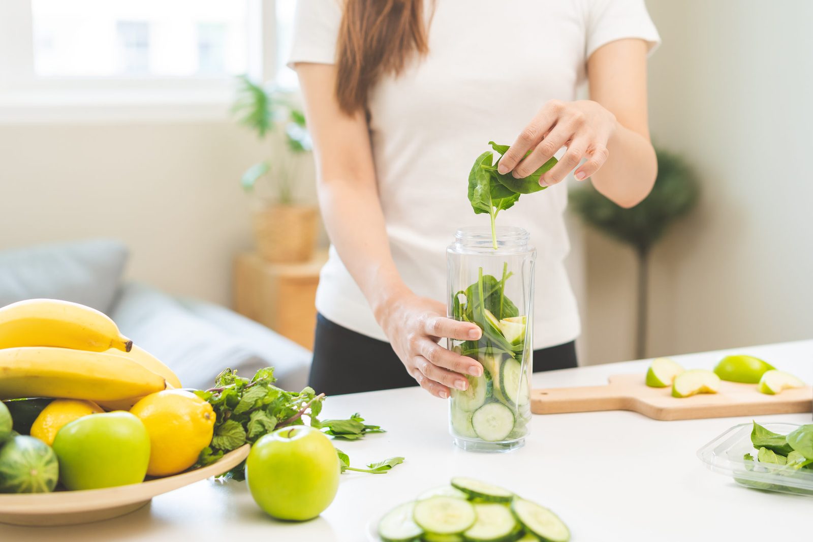 Eine Frau im weißen T-Shirt gibt frische Spinatblätter in einen Mixbecher, in dem schon Gurkenscheiben liegen; auf dem Küchentisch stehen Bananen, Zitronen, grüne Äpfel und ein Brett mit Apfelstücken.