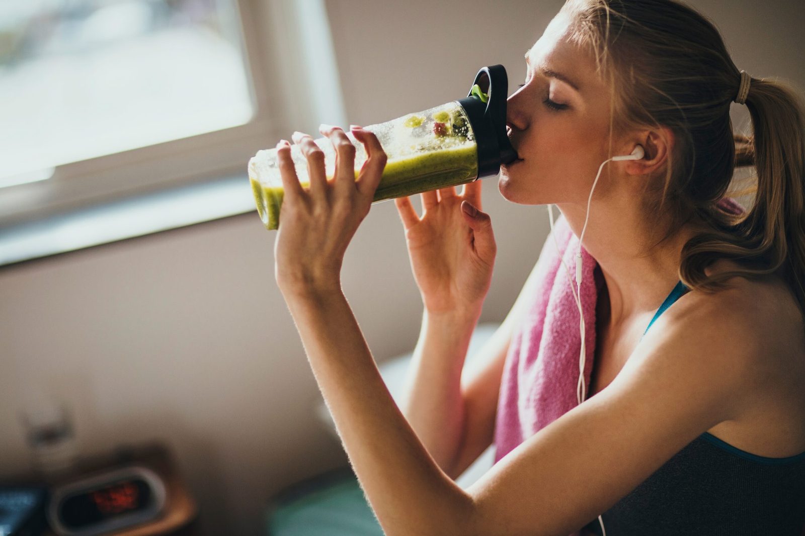 Junge Frau im Sporttop mit Handtuch über der Schulter trinkt nach dem Training zu Hause einen grünen Shake aus einem durchsichtigen Shaker, im Hintergrund ein helles Fenster.