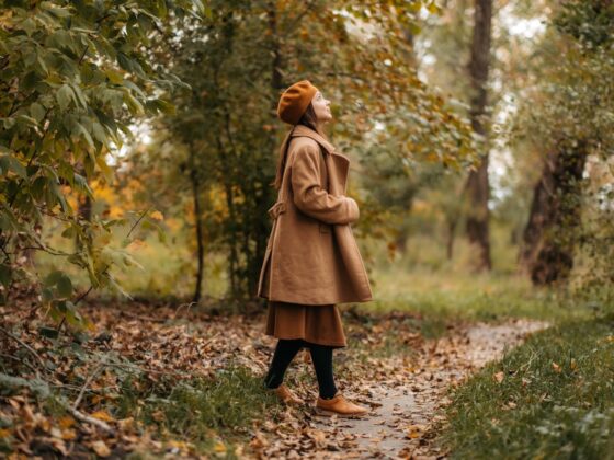 Frau in braunem Wollmantel steht seitlich auf einem laubbedeckten Weg im Herbstpark und schaut in die Baumkronen.