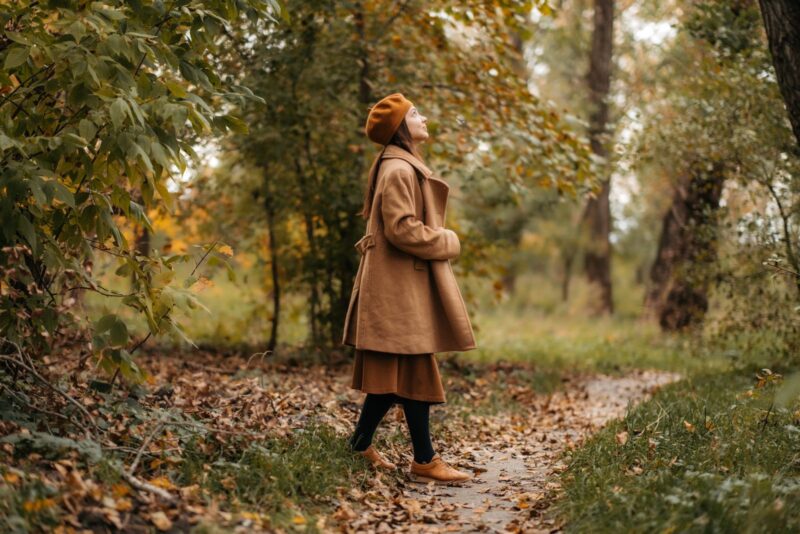 Frau in braunem Wollmantel steht seitlich auf einem laubbedeckten Weg im Herbstpark und schaut in die Baumkronen.