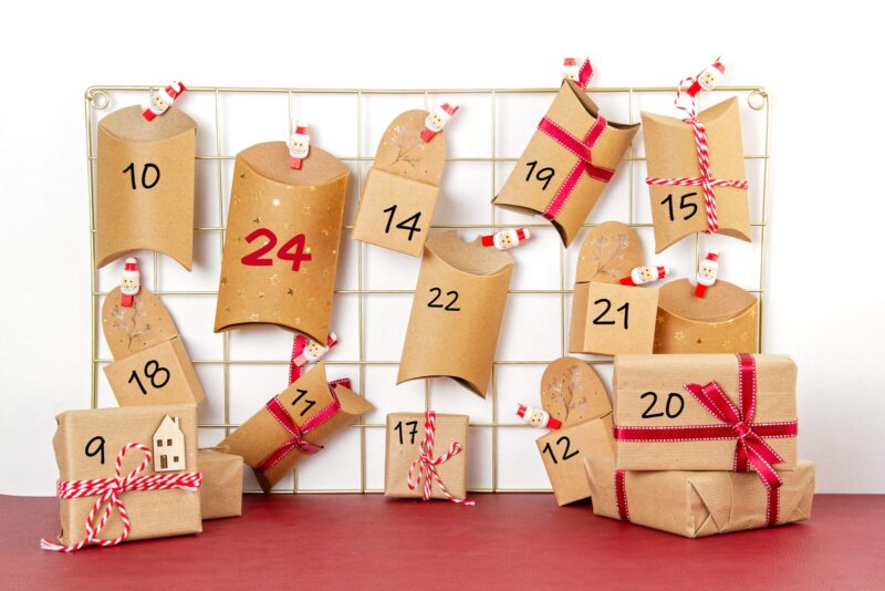 Selbstgemachter Adventskalender aus Kraftpapier-Schachteln mit Zahlen und roten Schleifen an einem Metallgitter